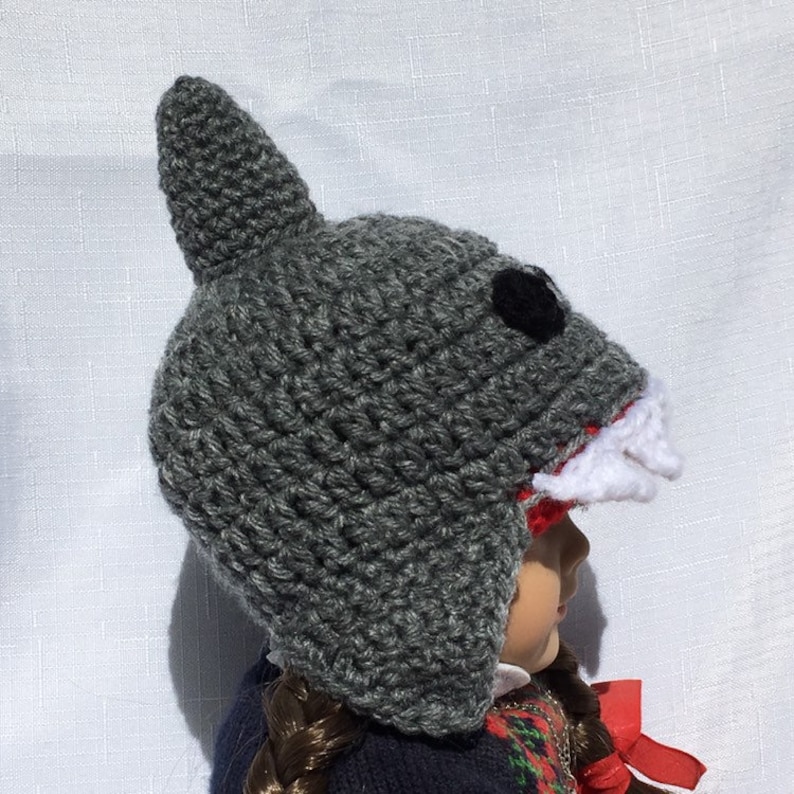 May include: Gray crocheted shark hat with a white and red mouth and black eyes. The hat is worn by a doll with brown hair.