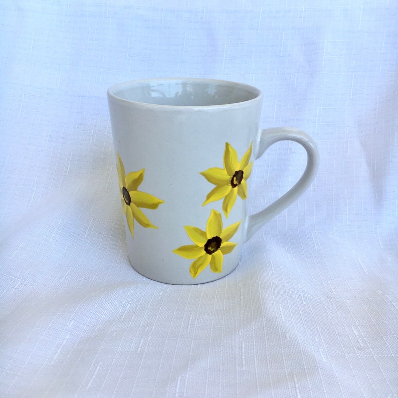 K&ouml;nnte beinhalten: Wei&szlig;e Keramiktasse mit gelbem und braunem Blumendesign. Die Tasse hat einen Henkel und ist mit vier gelben Blumen mit braunen Zentren verziert.