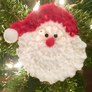 Peut inclure: Ornement de No&euml;l en crochet blanc repr&eacute;sentant un P&egrave;re No&euml;l avec un chapeau rouge et des yeux globuleux. L'ornement est suspendu &agrave; un sapin de No&euml;l.