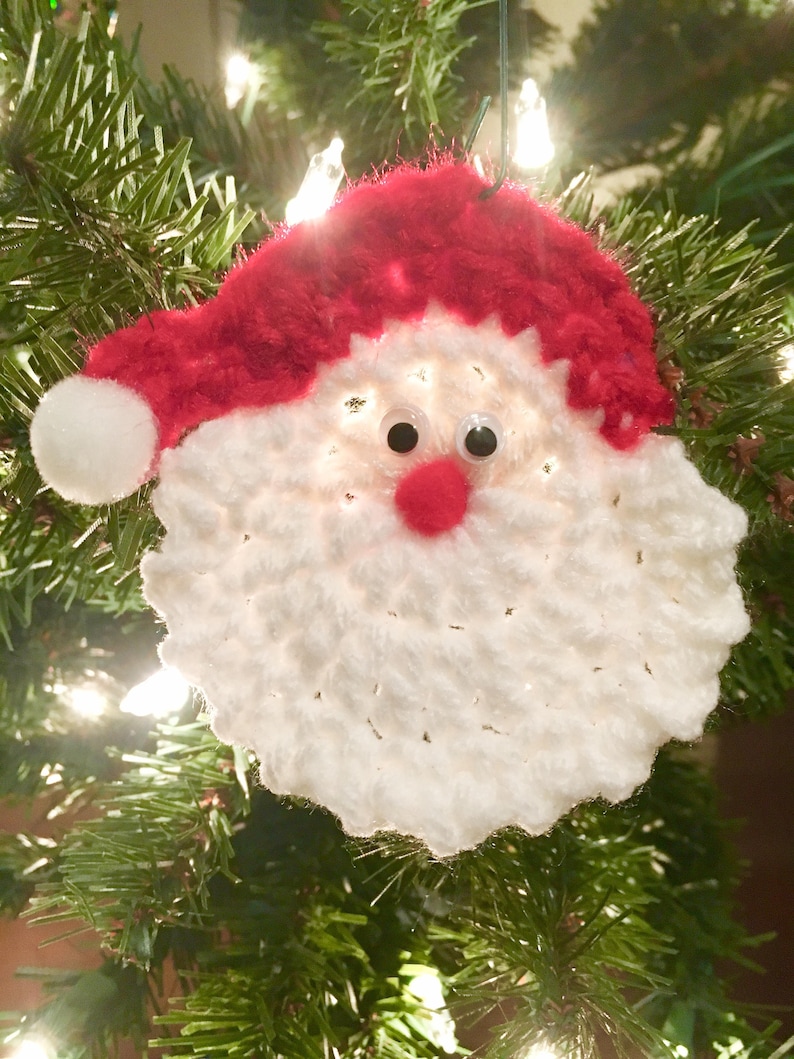 Peut inclure: Ornement de No&euml;l en crochet blanc repr&eacute;sentant un P&egrave;re No&euml;l avec un chapeau rouge et des yeux globuleux. L'ornement est suspendu &agrave; un sapin de No&euml;l.