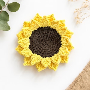 May include: A round, crocheted sunflower coaster featuring a dark brown circular center surrounded by two layers of bright yellow, pointed petals. The item is crafted with visible yarn stitches, creating a textured, floral-inspired design.