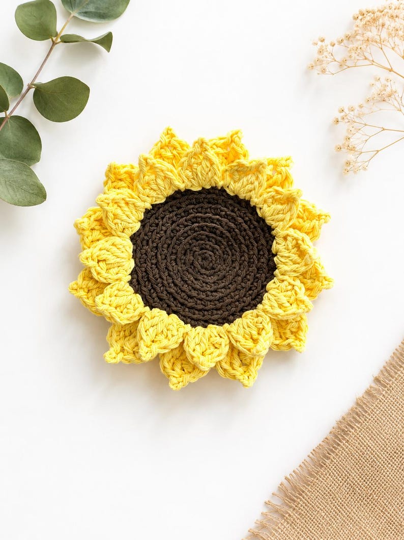 May include: A round, crocheted sunflower coaster featuring a dark brown circular center surrounded by two layers of bright yellow, pointed petals. The item is crafted with visible yarn stitches, creating a textured, floral-inspired design.