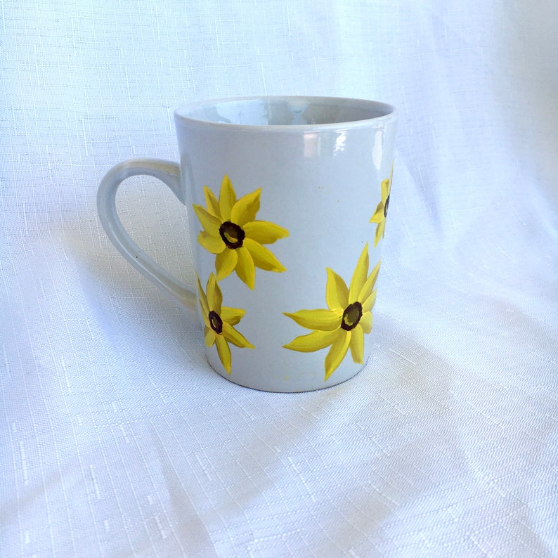 K&ouml;nnte beinhalten: Eine wei&szlig;e Keramiktasse mit Henkel, verziert mit gelben Blumen mit braunen Zentren. Die Tasse steht auf einem wei&szlig;en Hintergrund.