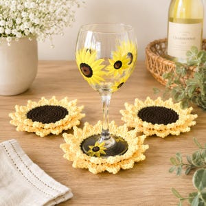 May include: A clear glass wine glass featuring hand-painted yellow sunflower designs with dark brown centers. The glass is surrounded by three round, crocheted sunflower coasters with dark brown centers and yellow petal-like edges.