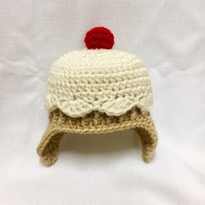 May include: A crocheted hat designed to resemble a cupcake, featuring a tan base, a cream-colored top with a scalloped edge, and a bright red pom-pom on the very top. The hat has two side flaps extending downward from the main body.