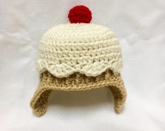 May include: A crocheted hat designed to resemble a cupcake, featuring a tan base, a cream-colored top with a scalloped edge, and a bright red pom-pom on the very top. The hat has two side flaps extending downward from the main body.
