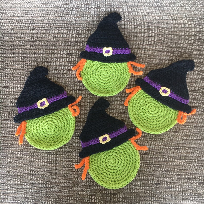 Op de afbeelding: Vier gehaakte heksenonderzetters met zwarte hoeden en groene bases. De onderzetters hebben een oranje franje rond de randen.