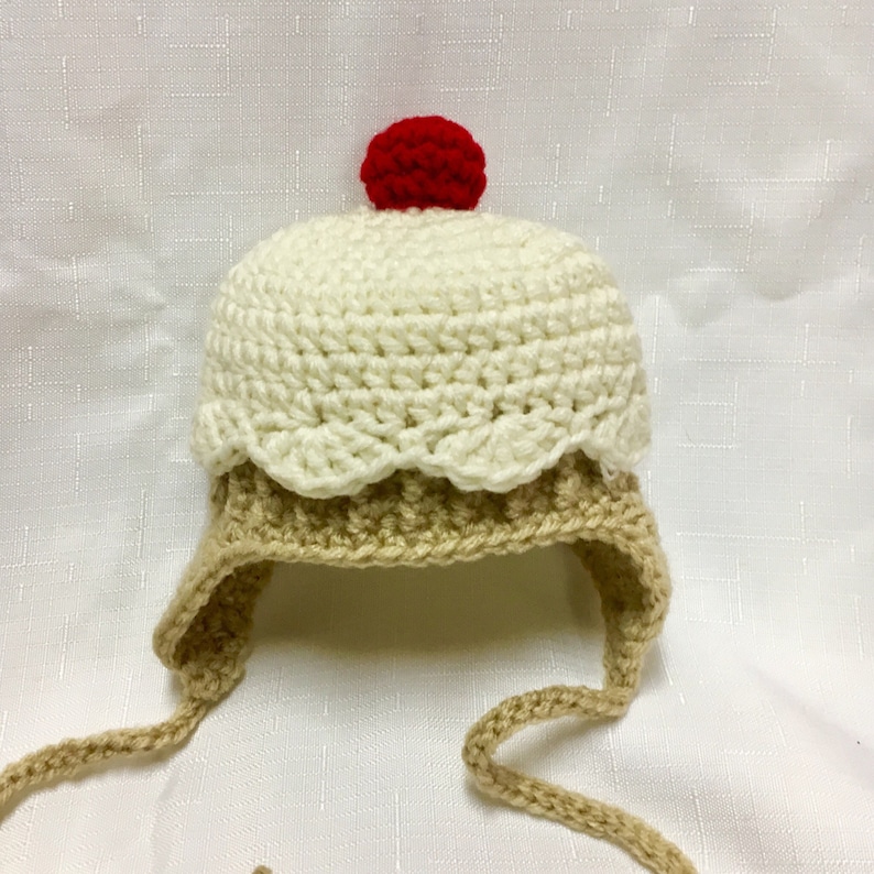 May include: A crocheted baby hat shaped like a cupcake with a red cherry on top. The hat is white and brown with a brown tie.
