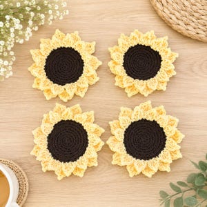 May include: Four round crochet coasters shaped like sunflowers, each featuring a dark brown circular center surrounded by layered, textured yellow petals. The coasters are arranged in a square formation, showcasing the intricate yarn stitching.