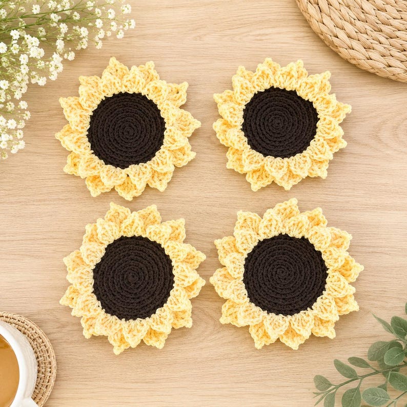 May include: Four round crochet coasters shaped like sunflowers, each featuring a dark brown circular center surrounded by layered, textured yellow petals. The coasters are arranged in a square formation, showcasing the intricate yarn stitching.