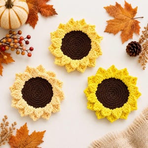 May include: Three round crochet coasters shaped like sunflowers, each featuring a dark brown center and yellow or light yellow petals. The coasters are arranged on a surface surrounded by autumn leaves, a small pumpkin, and a pinecone.