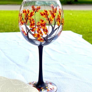 May include: A clear glass wine goblet featuring a hand-painted design of autumn tree branches with vibrant red, orange, and yellow leaves. The stem and base are painted solid black, with additional small colorful leaf accents on the base.