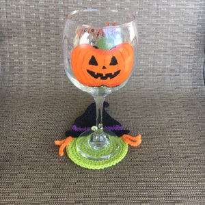 Op de afbeelding: Een glas met een geschilderde oranje pompoen met een jack-o'-lantern gezicht erop. Het glas staat op een groene en zwarte gehaakte heksenhoed onderzetter.