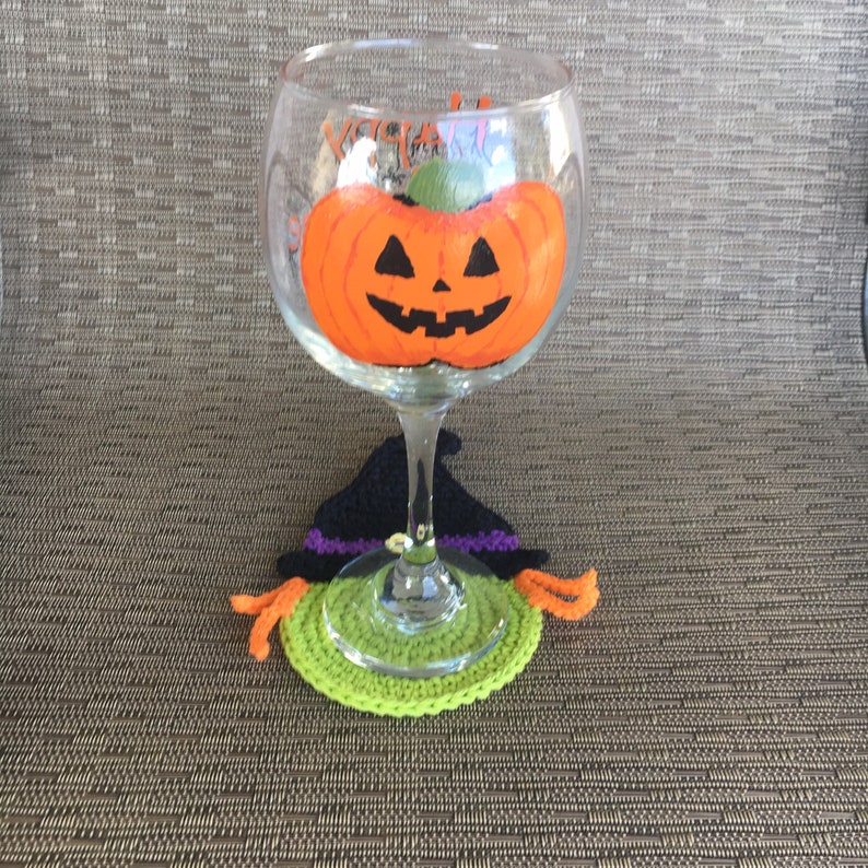 Op de afbeelding: Een glas met een geschilderde oranje pompoen met een jack-o'-lantern gezicht erop. Het glas staat op een groene en zwarte gehaakte heksenhoed onderzetter.