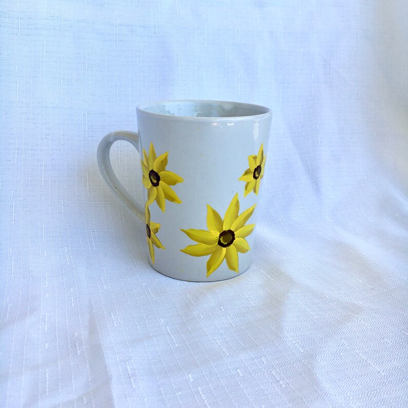 K&ouml;nnte beinhalten: Wei&szlig;e Keramiktasse mit Henkel, verziert mit einem handgemalten Design aus gelben Blumen mit braunen Zentren.