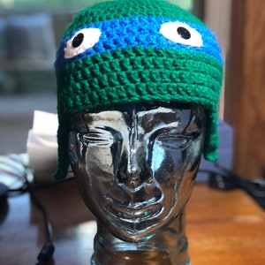 May include: Green crocheted hat with blue stripes and white googly eyes. The hat is on a glass head form.