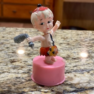 May include: A vintage plastic finger puppet of a cartoon caveman with a club. The puppet is wearing a red and brown loincloth and has a pink base.