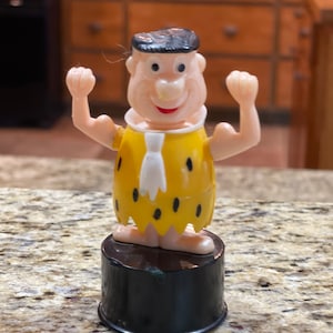 May include: A yellow and black plastic figurine of a cartoon character with a white necktie, standing on a black base. The character is wearing a yellow loincloth with black spots and has a black hair.