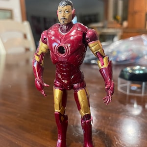 May include: A red and gold Iron Man action figure. The figure has a detailed face and is wearing a red and gold suit. The figure is standing on a wooden surface.