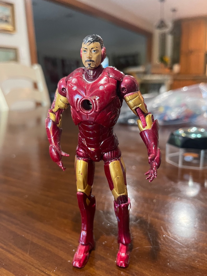 May include: A red and gold Iron Man action figure. The figure has a detailed face and is wearing a red and gold suit. The figure is standing on a wooden surface.