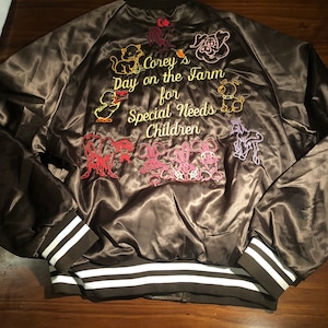 May include: A brown satin bomber jacket with white stripes on the cuffs and waistband. The back of the jacket features colorful embroidered animals and the text "Corey's Day on the Farm for Special Needs Children."