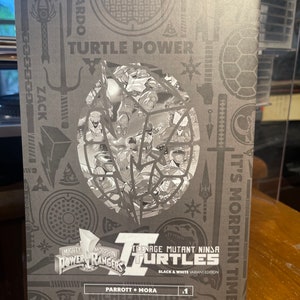 May include: A black and white comic book cover featuring the Teenage Mutant Ninja Turtles and the Mighty Morphin Power Rangers. The cover has the title "Teenage Mutant Ninja Turtles" and "Black & White Variant Edition". The cover also has the text "It's Morphin Time" and "Parrott &bull; Mora #1".