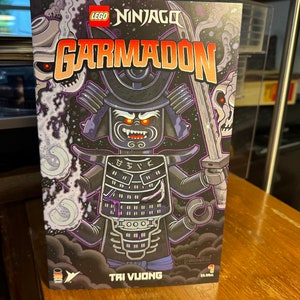 May include: A comic book cover featuring a black and white illustration of a Lego Ninjago character, Garmadon, in a samurai suit. The background is a dark blue and purple space scene with white clouds. The title of the comic is "LEGO Ninjago Garmadon".