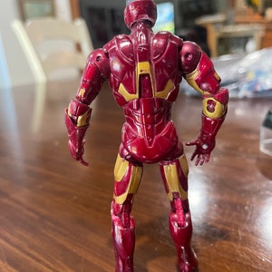 May include: A red and gold action figure of a superhero, viewed from the back. The figure has a metallic appearance with gold accents on the arms, legs, and back. The figure is standing on a wooden surface.