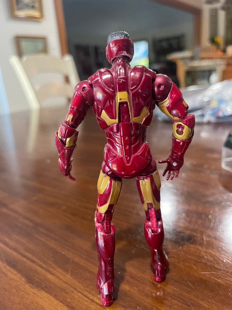 May include: A red and gold action figure of a superhero, viewed from the back. The figure has a metallic appearance with gold accents on the arms, legs, and back. The figure is standing on a wooden surface.