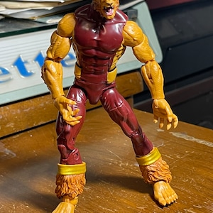 May include: A red and gold action figure of a werewolf with a snarling expression. The figure has a muscular build and is posed in a dynamic stance.