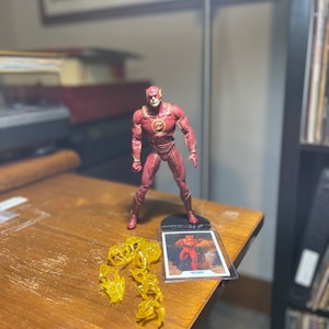 May include: A red and gold action figure of The Flash, a superhero with super speed, standing on a wooden table. The figure is holding a lightning bolt in its hand. There is a yellow plastic lightning bolt effect on the table in front of the figure. The figure is also featured on a trading card with the text "The Flash" on it.
