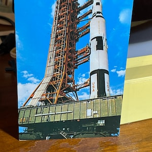 May include: A vintage postcard featuring a black and white rocket on a launchpad. The rocket is labeled "USA" and has a red, white, and blue color scheme. The launchpad is made of metal and has a gray color.
