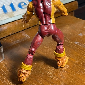 May include: A red and yellow action figure of a superhero with a furry head and tail. The figure is posed in a standing position with its arms raised. The figure has a hole in its back.