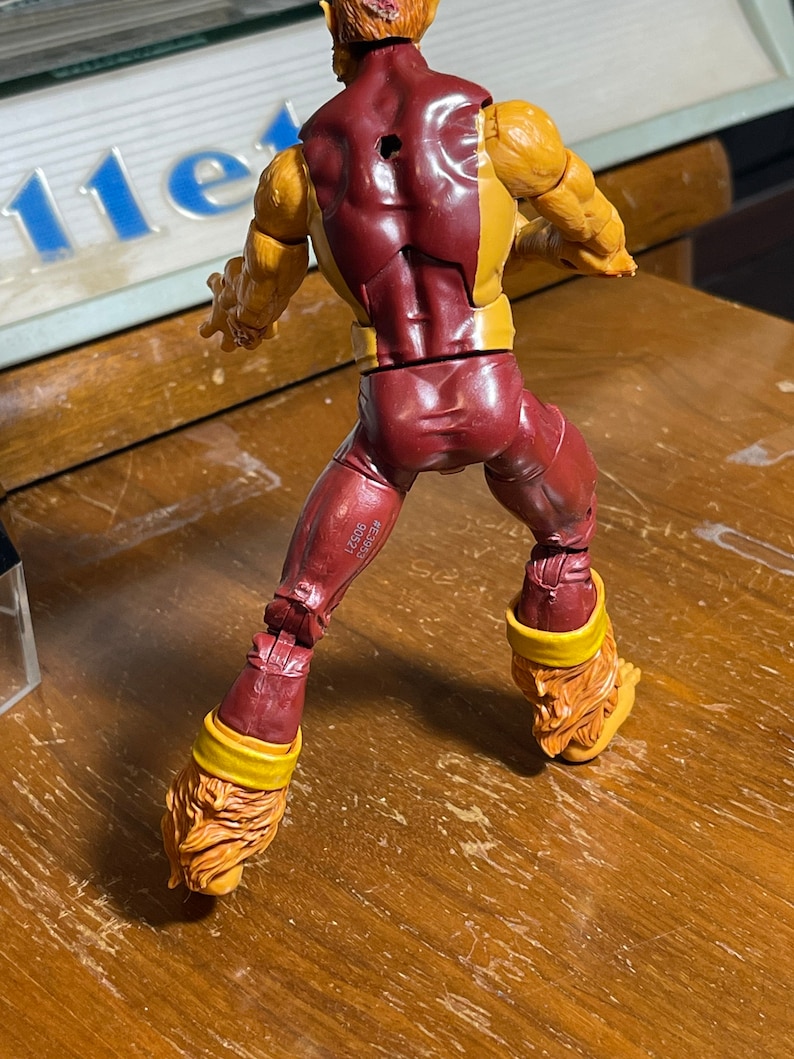 May include: A red and yellow action figure of a superhero with a furry head and tail. The figure is posed in a standing position with its arms raised. The figure has a hole in its back.