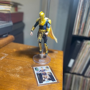 May include: A gold and blue action figure of a superhero wearing a helmet and cape. The figure is standing on a clear plastic base with a DC Comics logo. The figure is holding a card with the text "Mr.  Mind" on it.