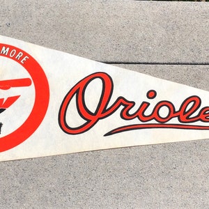 May include: A vintage pennant in a triangular shape, featuring the Baltimore Orioles logo. The pennant is primarily white with orange and black accents. The word "Orioles" is written in a stylized script.