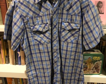 Pode incluir: Uma camisa de manga curta xadrez azul e cinza com uma etiqueta Wrangler. A camisa tem dois bolsos no peito com fecho de botão.