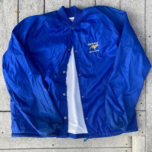 May include: A blue nylon jacket with a snap closure and a white lining. The jacket has an embroidered logo that says "Exxon-West Safety Around".