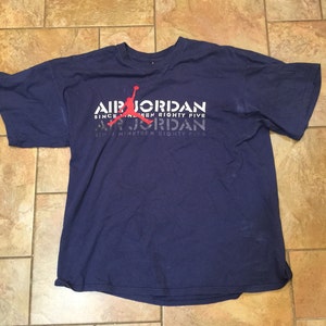 May include: A navy blue t-shirt with a white graphic print of a basketball player with the text "Air Jordan Since Nineteen Eighty Five" repeated twice.
