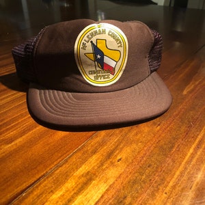May include: A brown mesh trucker hat with a yellow patch featuring a Texas flag and the text "McLennan County Constable Office."