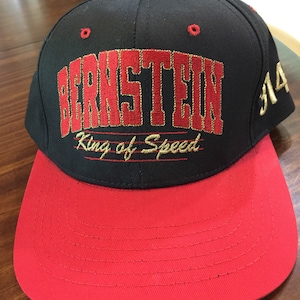 May include: Black and red baseball cap with the name "BERNSTEIN" embroidered in red with gold trim. The text "King of Speed" is below the name. The brim is red, and the top of the cap is black. The number "14" is embroidered in gold on the side.