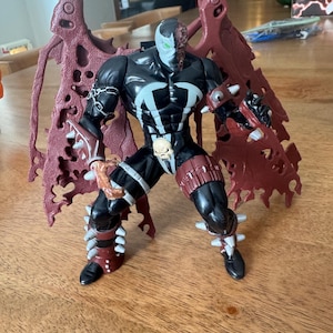 May include: A Spawn action figure in a dynamic pose, featuring a black and white costume with a skull emblem. The figure has a red cape and spiked accents on the boots and gauntlets. The figure is approximately 7 inches tall.
