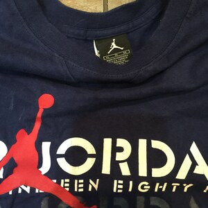 May include: A navy blue t-shirt with a red silhouette of a basketball player dunking. The text "JORDAN" is printed in white on the shirt, with the words "NINE-EIGHTY" printed in white below it.