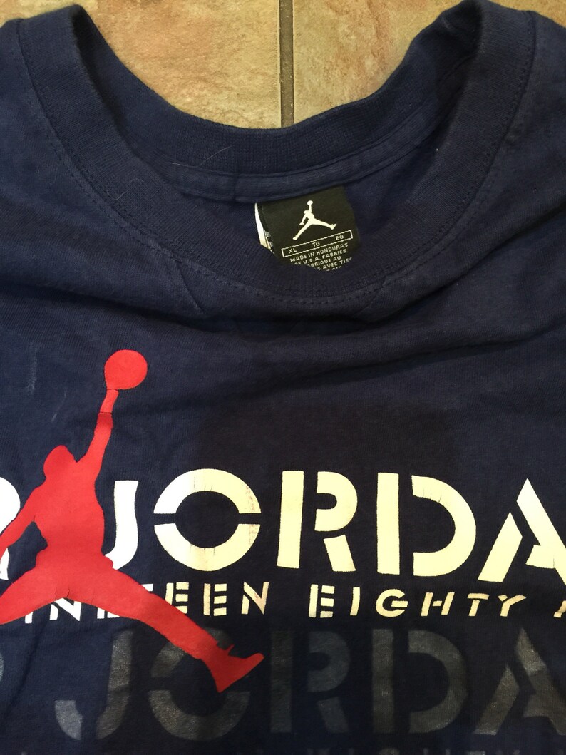 May include: A navy blue t-shirt with a red silhouette of a basketball player dunking. The text "JORDAN" is printed in white on the shirt, with the words "NINE-EIGHTY" printed in white below it.