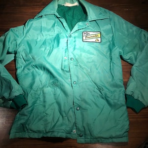 May include: A green jacket with a Paymaster logo patch on the chest. The jacket has a button-up front and two pockets.