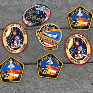 May include: Nine embroidered patches featuring the names of astronauts and space shuttle missions. The patches are yellow, red, blue, and white with black outlines. The patches are on a gray textured surface.