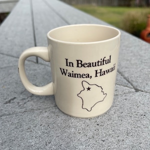 May include: A white ceramic coffee mug with a brown handle. The mug has the text "In Beautiful Waimea, Hawaii" and an outline of the island of Hawaii with a star on it.