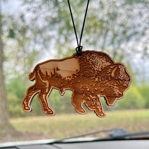May include: A wooden car air freshener shaped like a bison. The bison is brown and has a detailed design.