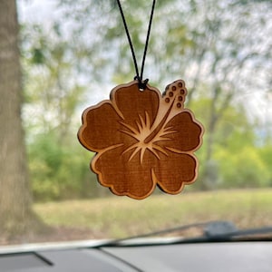 May include: A wooden car air freshener shaped like a brown hibiscus flower. The flower has a detailed design and is hanging from a black string.