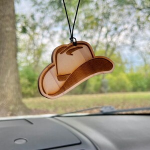 May include: A wooden cowboy hat car air freshener hanging from a rearview mirror. The hat is brown and has a simple design.